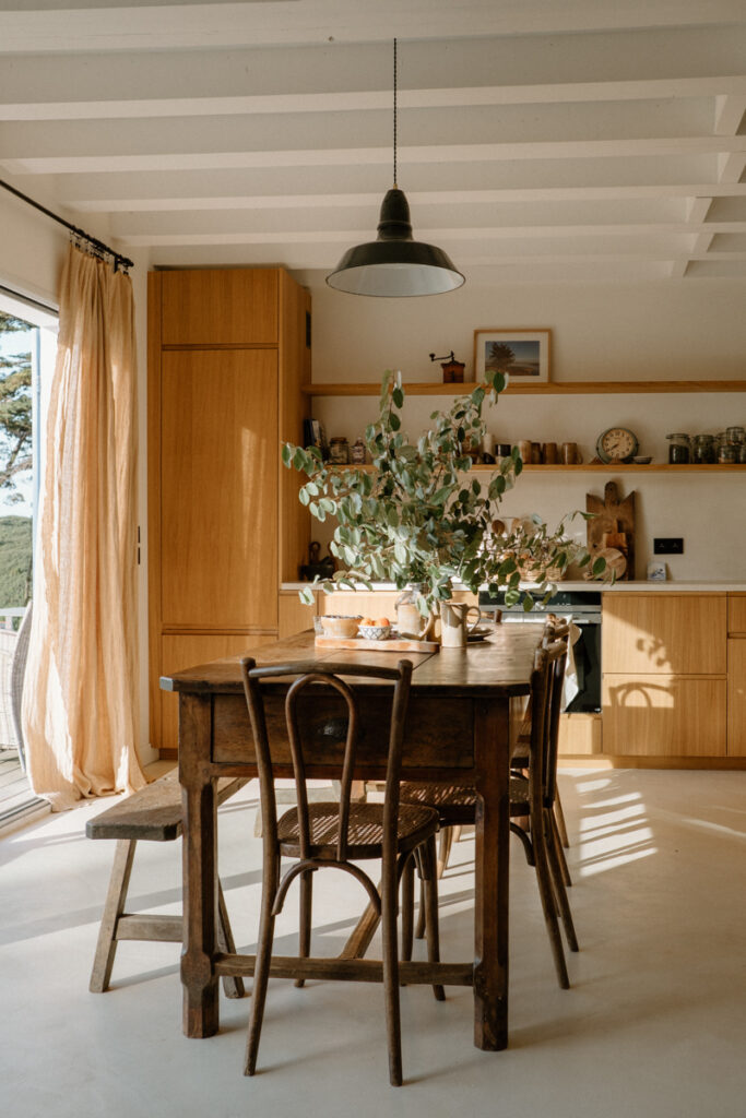 cuisine-dans-une-maison-secondaire-facades-bocklip-en-chene-naturel-30-10e-forme-moderne-grande-table-en-bois-au-centre-chaises-et-banc-autour-poutres-au-plafond-apparentes-etageres-murales-en-bois-avec-de-la-vaisselle-et-des-decorations