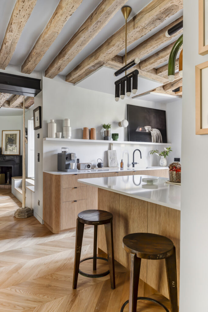 Cuisine renovée contemporaine avec façades Bocklip en chêne naturel plissé, plan de travail blanc mat, poutres apparentes et îlot central chaleureux.