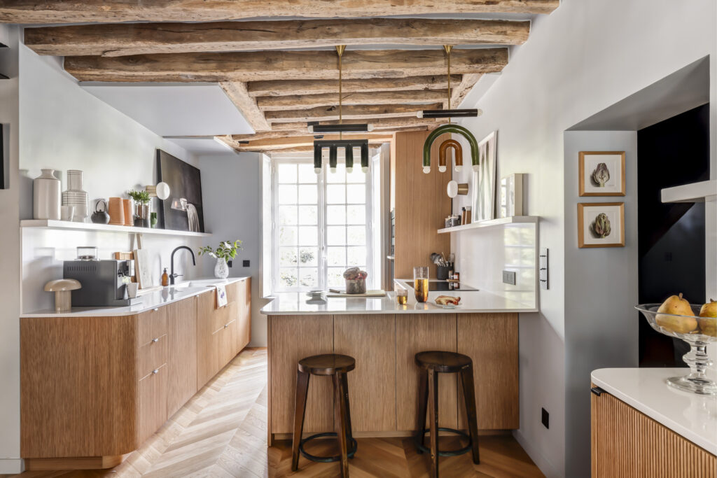 Cuisine rénovée contemporaine avec façades Bocklip en chêne naturel plissé, plan de travail blanc mat, poutres apparentes et îlot central chaleureux.